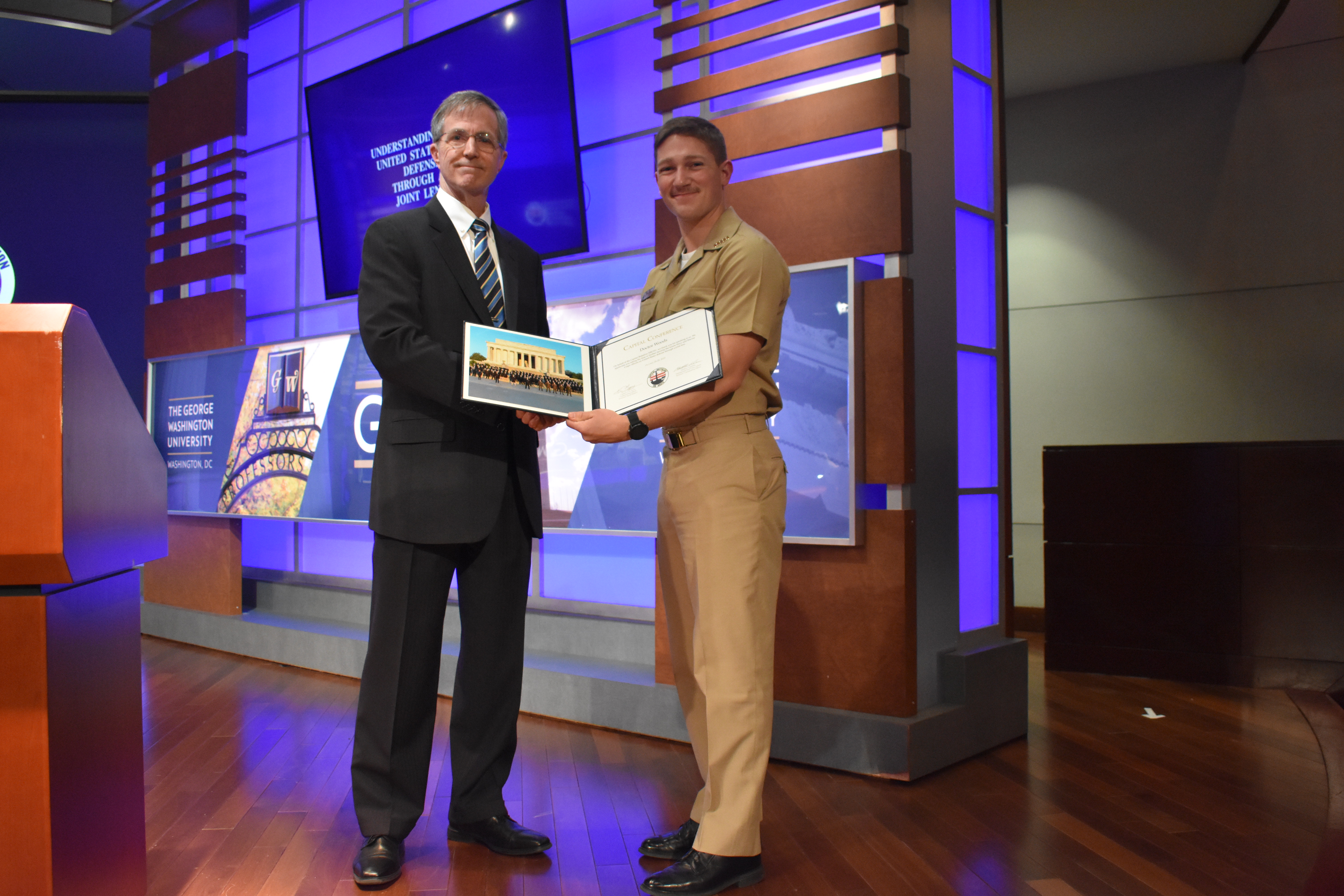 Two men stand on a stage. One is holding a certificate for the Capital Conference